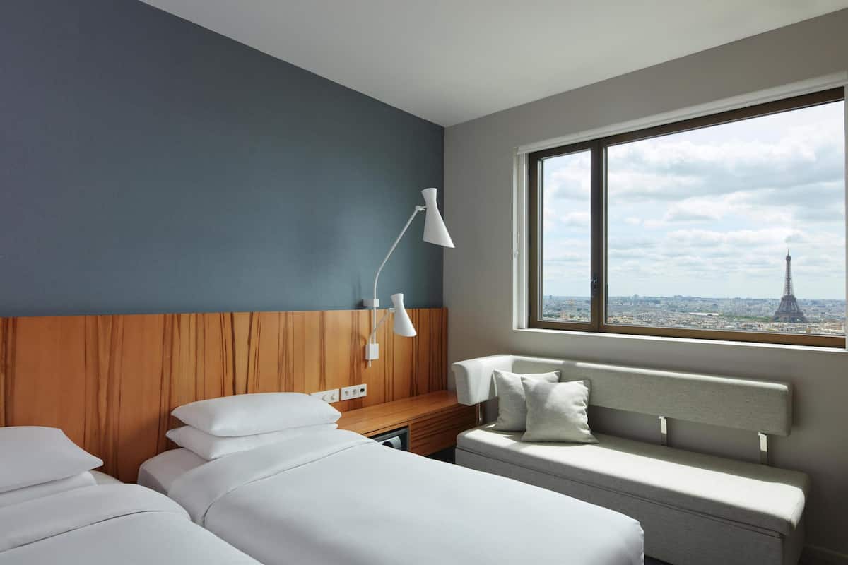 Hotellihuoneessa on sänky, penkki, työpöytä ja lamppu. Huoneesta on ikkuna, josta avautuu näkymä kaupungin siluettiin ja torniin.