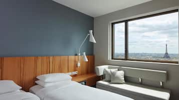 Habitación de hotel con cama, un banco, escritorio y lámpara. Se ve una ventana con vista a un paisaje urbano y una torre.