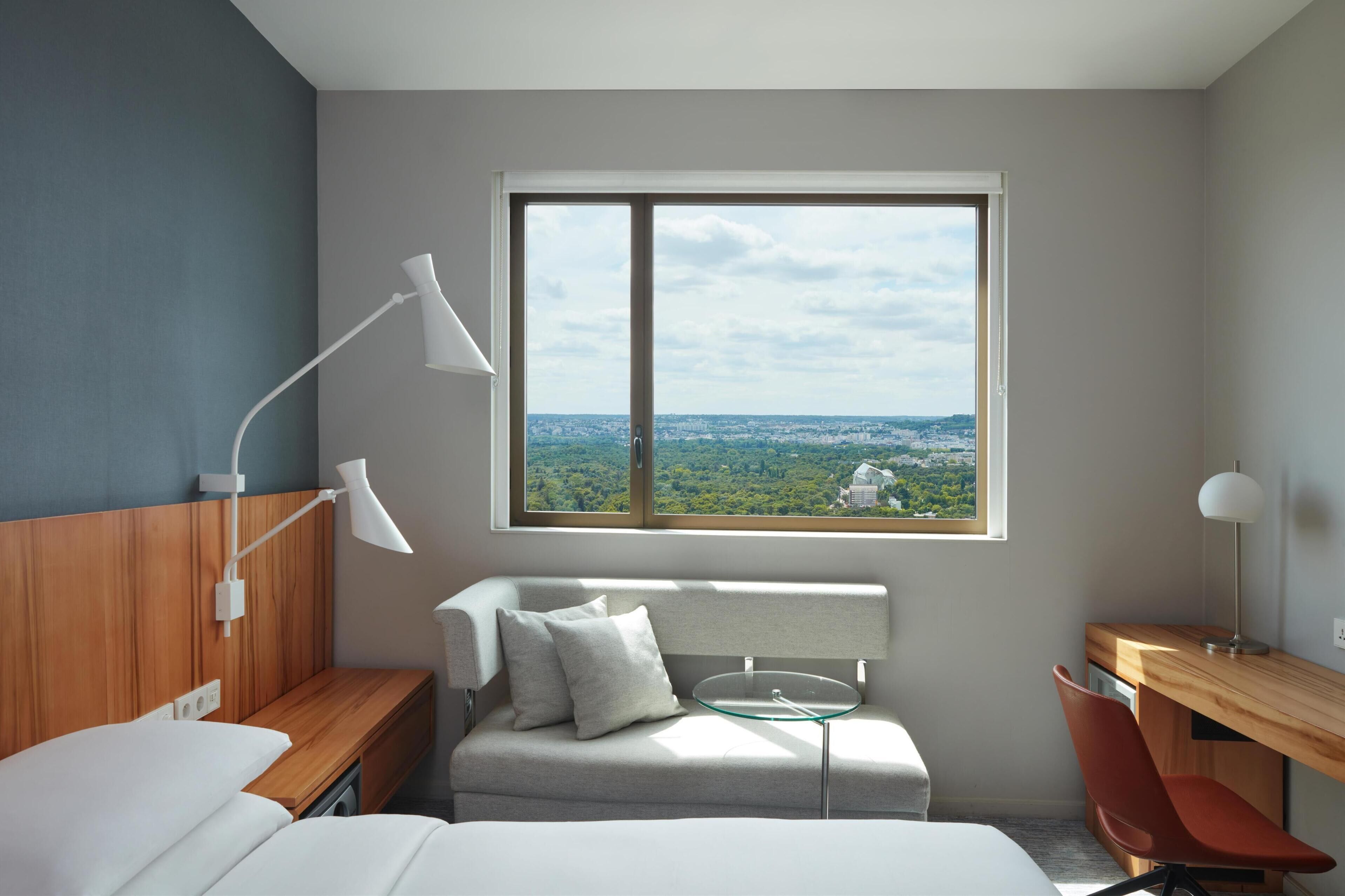 A modern hotel room with a bed, a desk, a chair, a window with a city view, and a lamp.