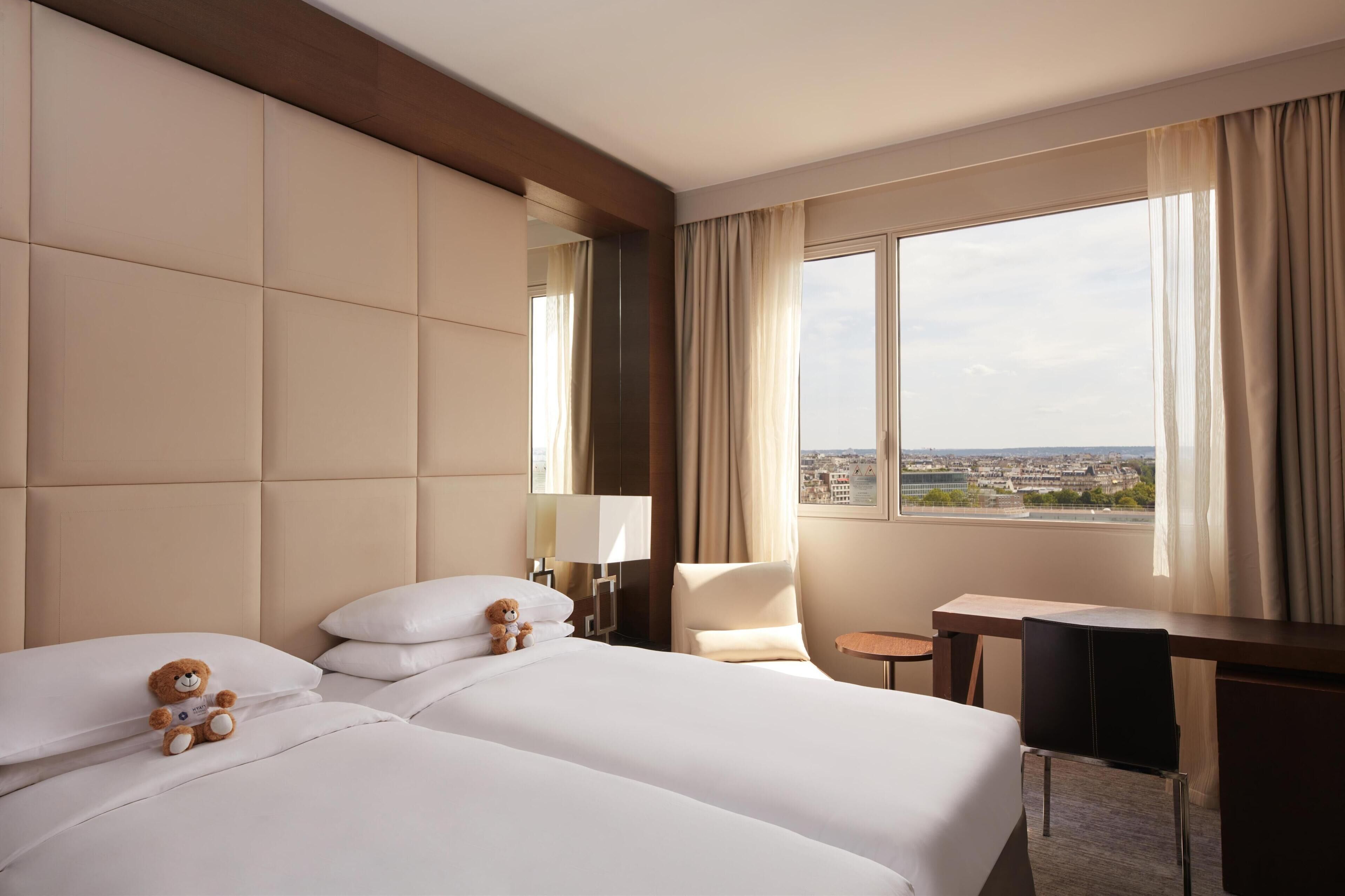 A hotel room with two beds, a desk, a chair, and a large window with a city view.
