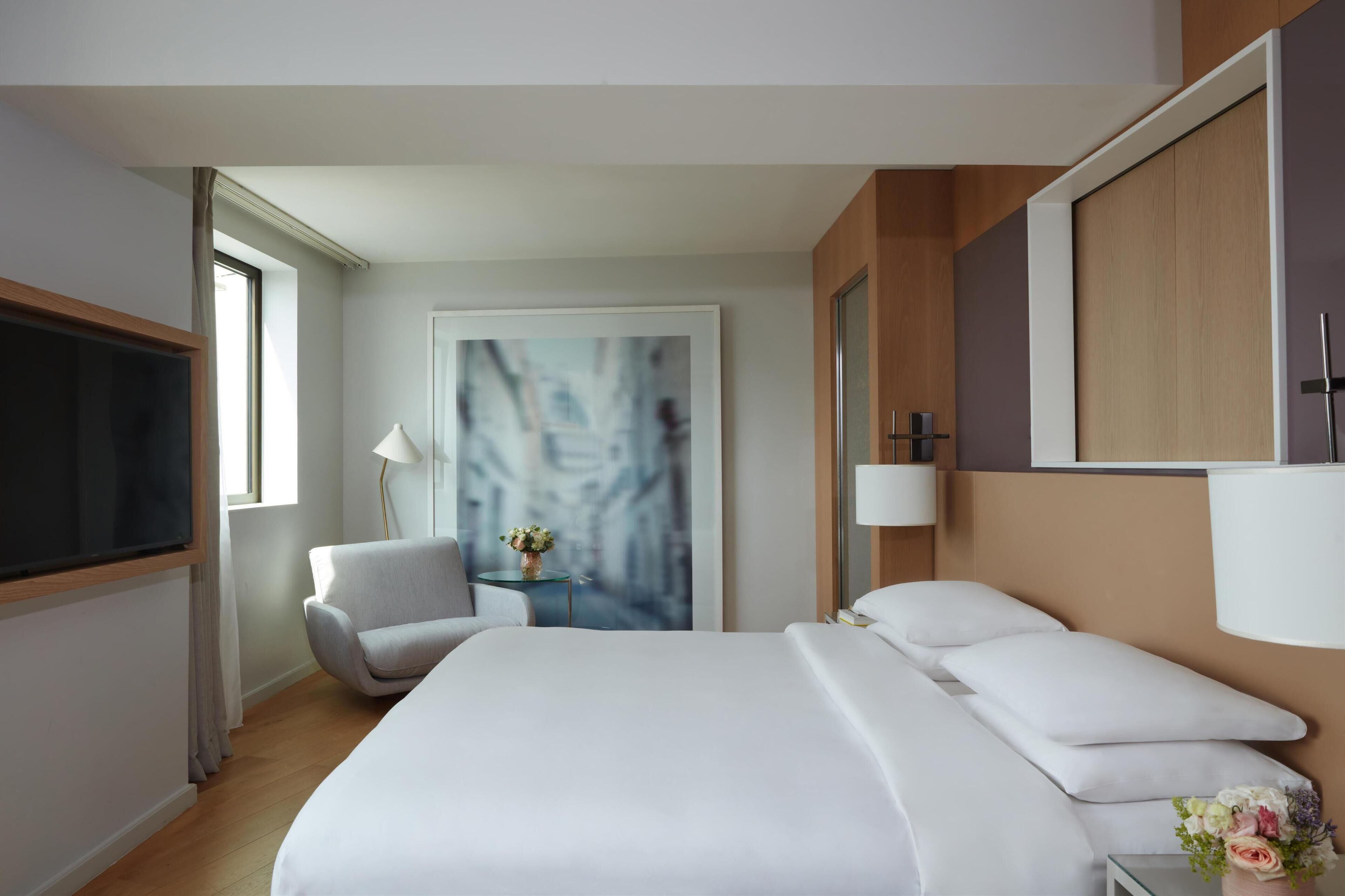 A modern hotel room with a large bed, a sitting area with a chair and table, a large window, and a painting on the wall.