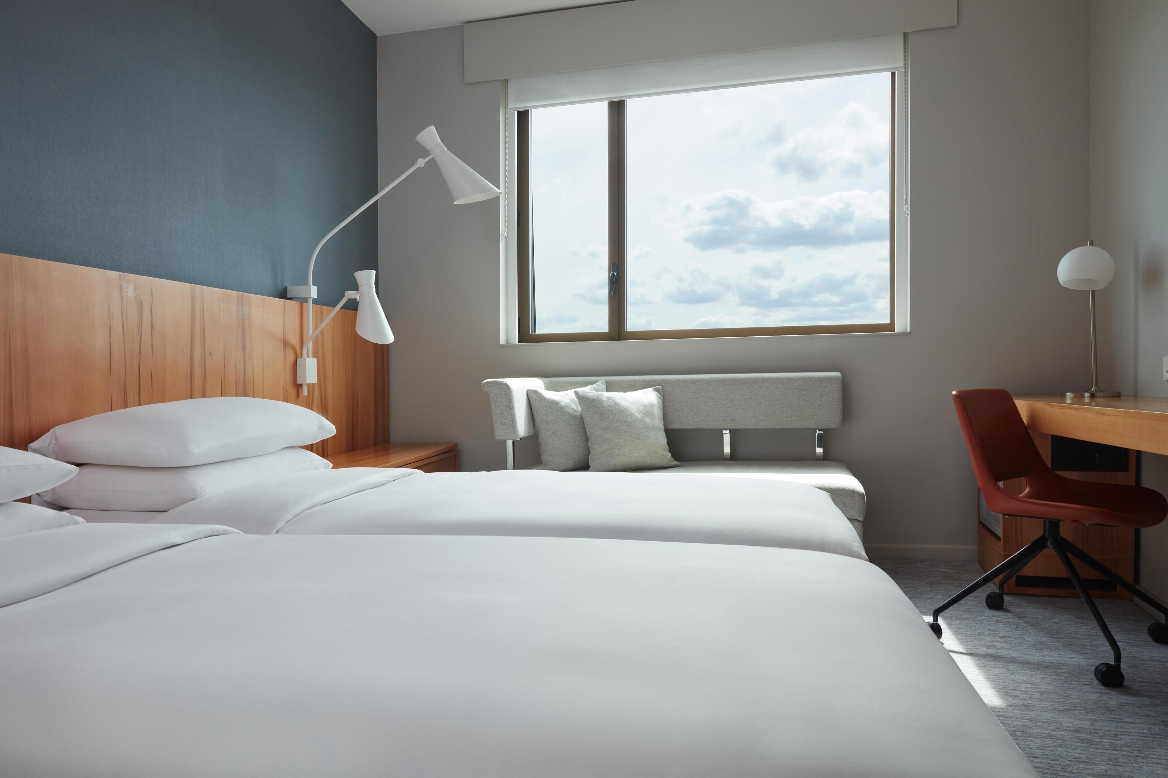 Habitación de hotel con dos camas, un escritorio, una silla y un ventanal con vistas al cielo.