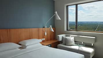 A hotel room with a large window, a bed, a sofa, a desk, and a lamp.