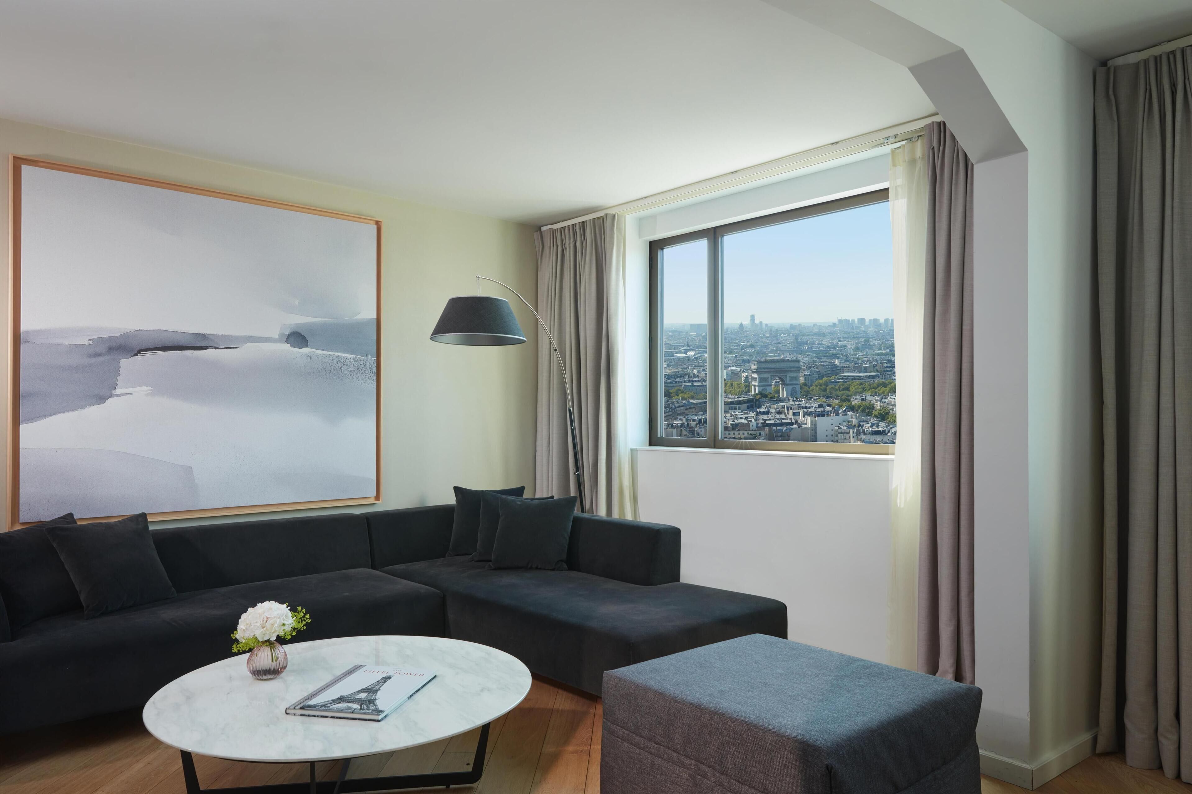 Une chambre d’hôtel moderne dotée d’une grande fenêtre donnant sur la ville, d’un canapé d’angle noir, d’une table basse ronde et d’un lampadaire.