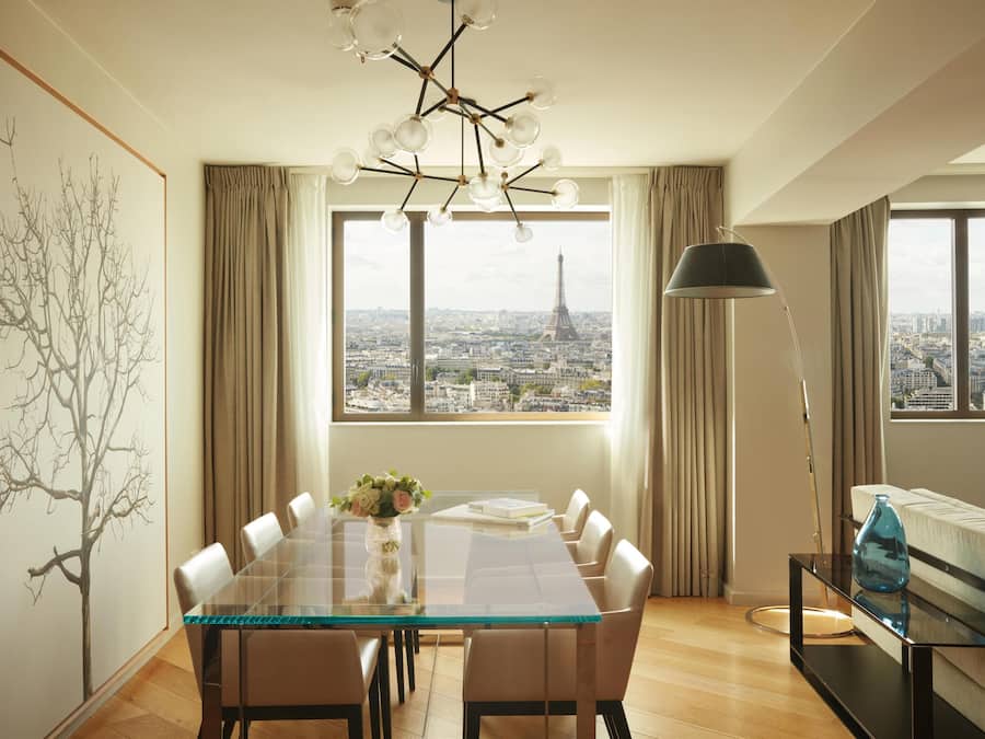 A dining area with a glass table, chairs, a vase of flowers, and a view of the Eiffel Tower through large windows.