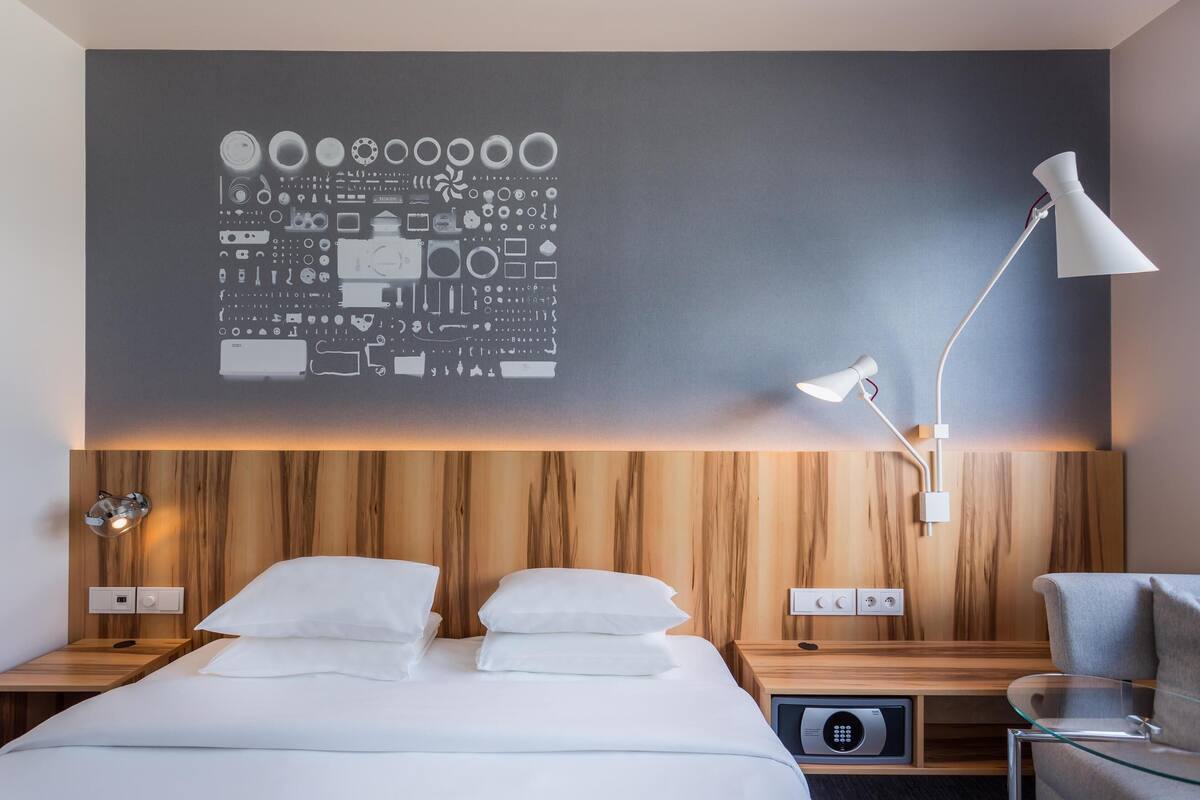 Moderni hotellihuone, jossa on puinen sängynpääty, suuri taideteos seinällä, yöpöytä lampun kanssa ja lasinen sohvapöytä.