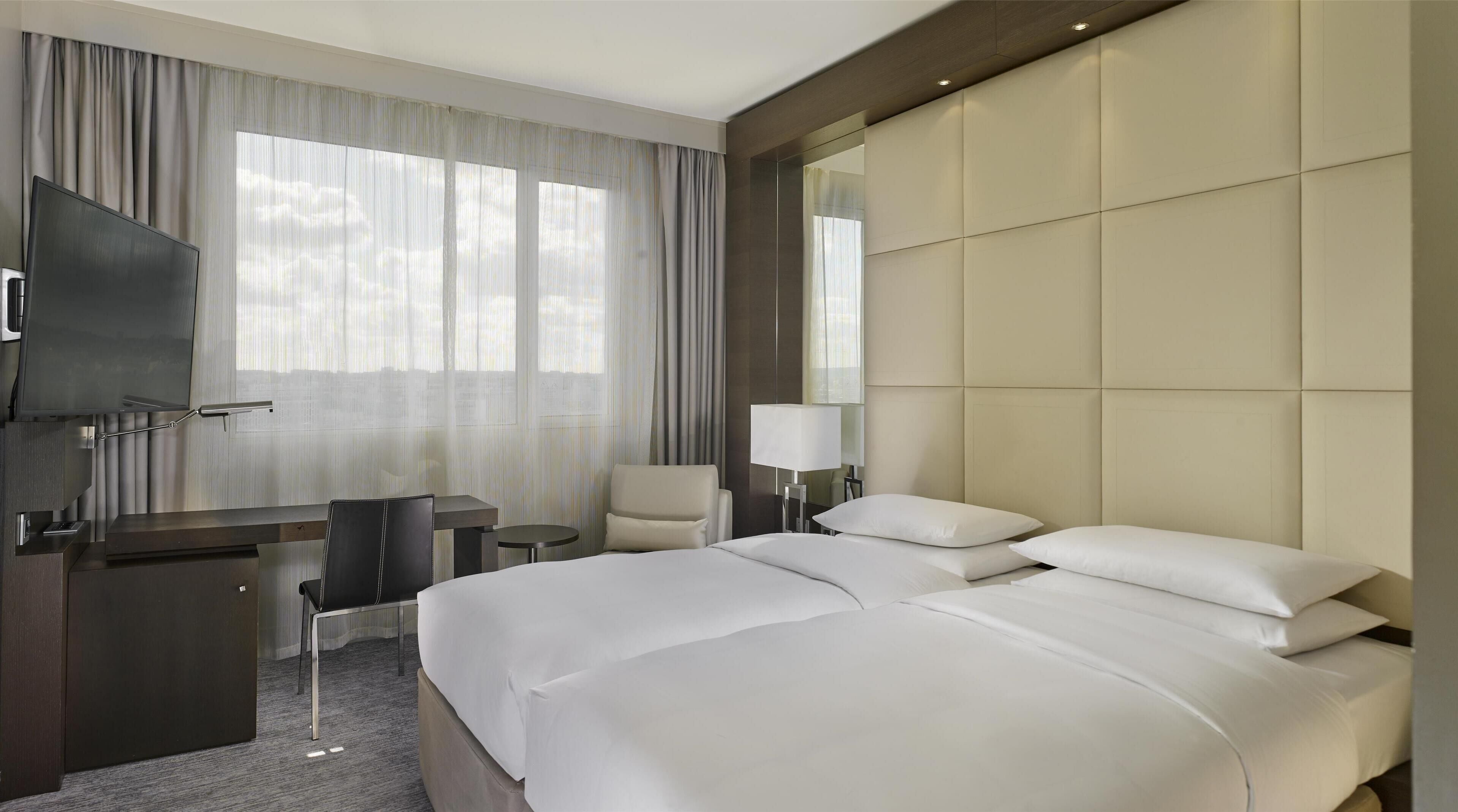 A hotel room with a large bed, a desk with a chair, a TV, and a large window with curtains.