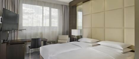 A hotel room with a large bed, a desk with a chair, a TV, and a large window with curtains.