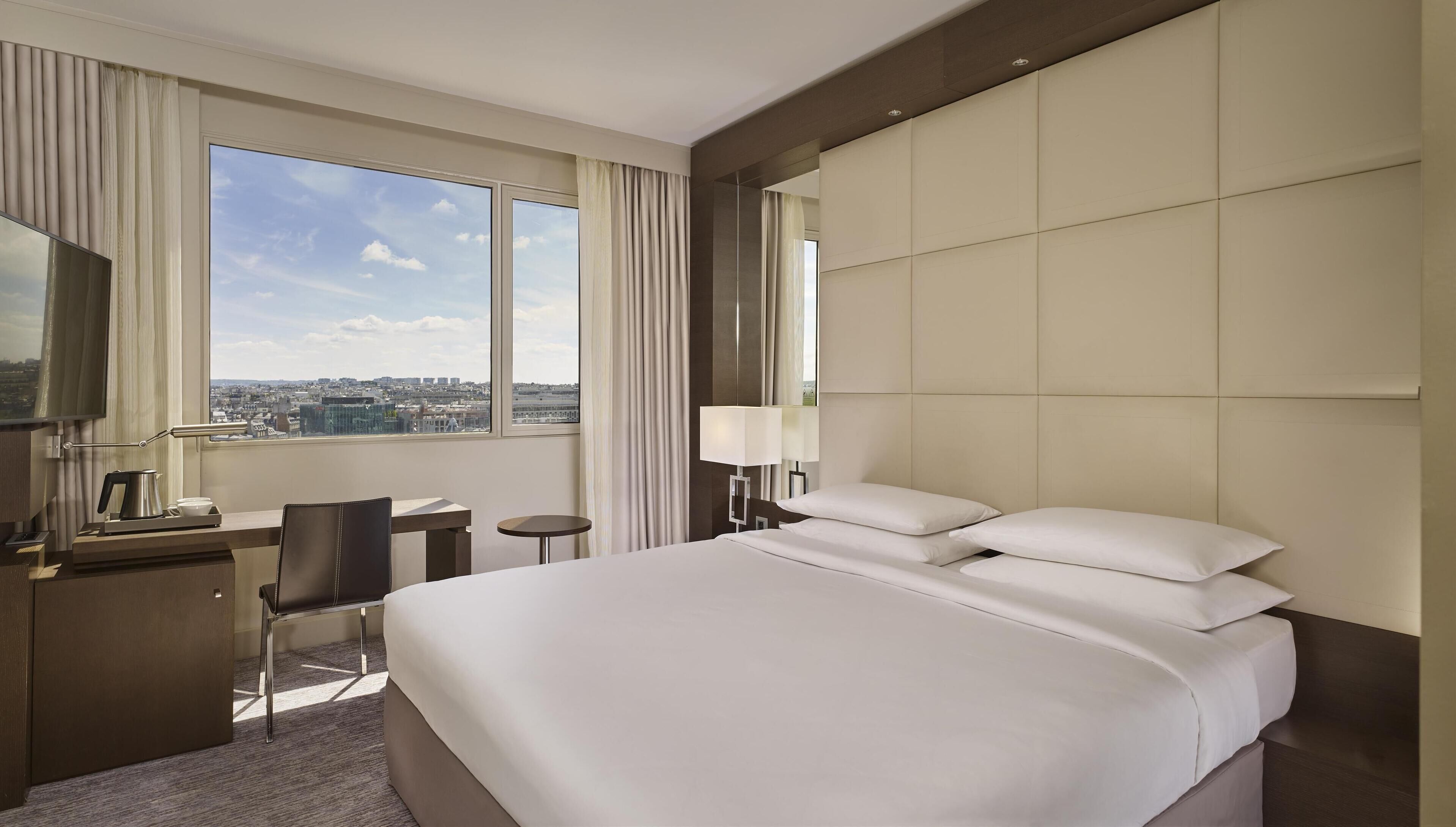A hotel room with a large bed, a desk with a chair, a small table, a lamp, and a TV.