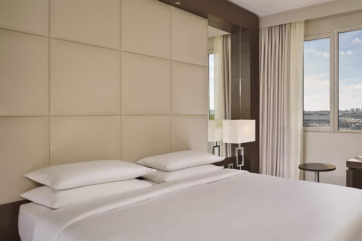 Moderni hotellihuone, jossa on suuri sänky, yöpöytä, lamppu ja ikkunasta avautuva näkymä kaupunkiin.