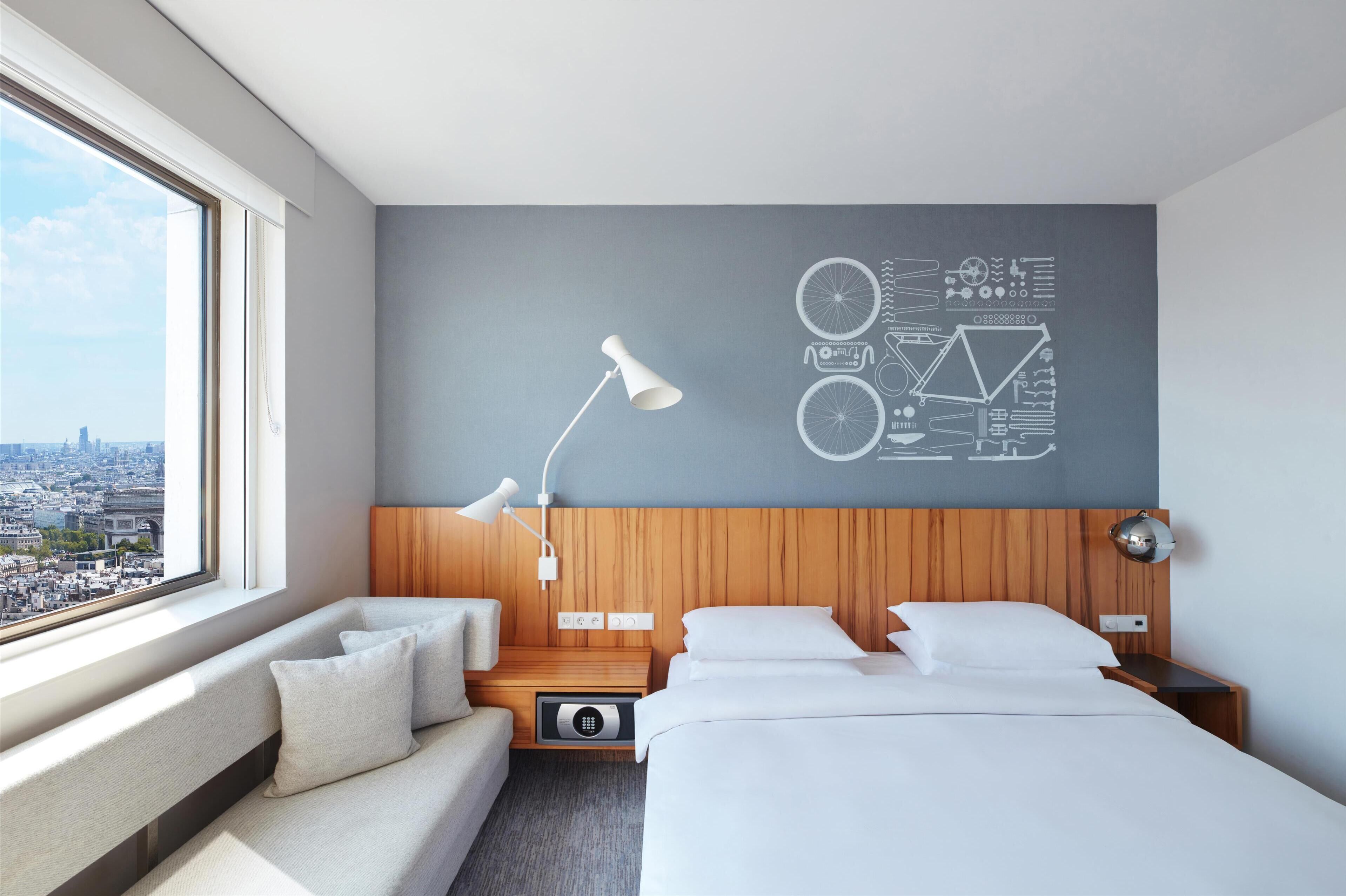 A modern hotel room with a city view, a large window, a bed, a sofa, and a wall with a bicycle-themed design.