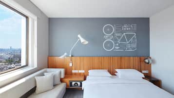 A modern hotel room with a city view, a large window, a bed, a sofa, and a wall with a bicycle-themed design.