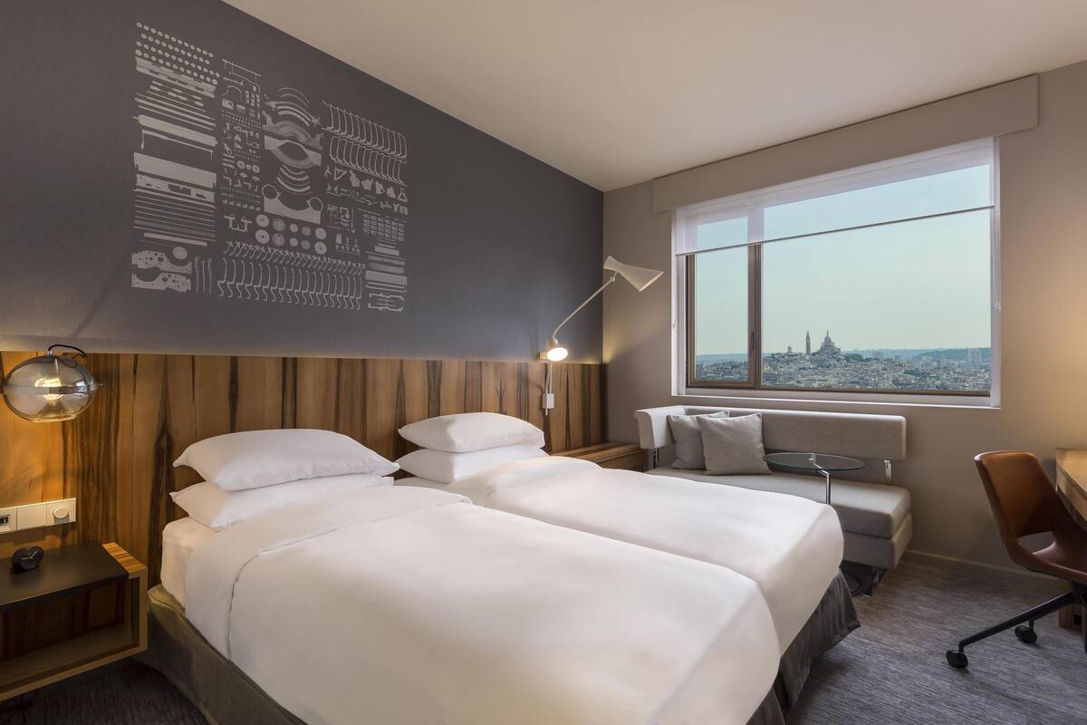 Hotellihuone, jossa on kaksi sänkyä, työpöytä, tuoli, lamppu ja suuri ikkuna, josta on näkymä kaupunkiin.