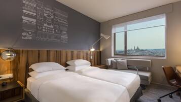 A hotel room with two beds, a desk, a chair, a lamp, and a large window with a city view.