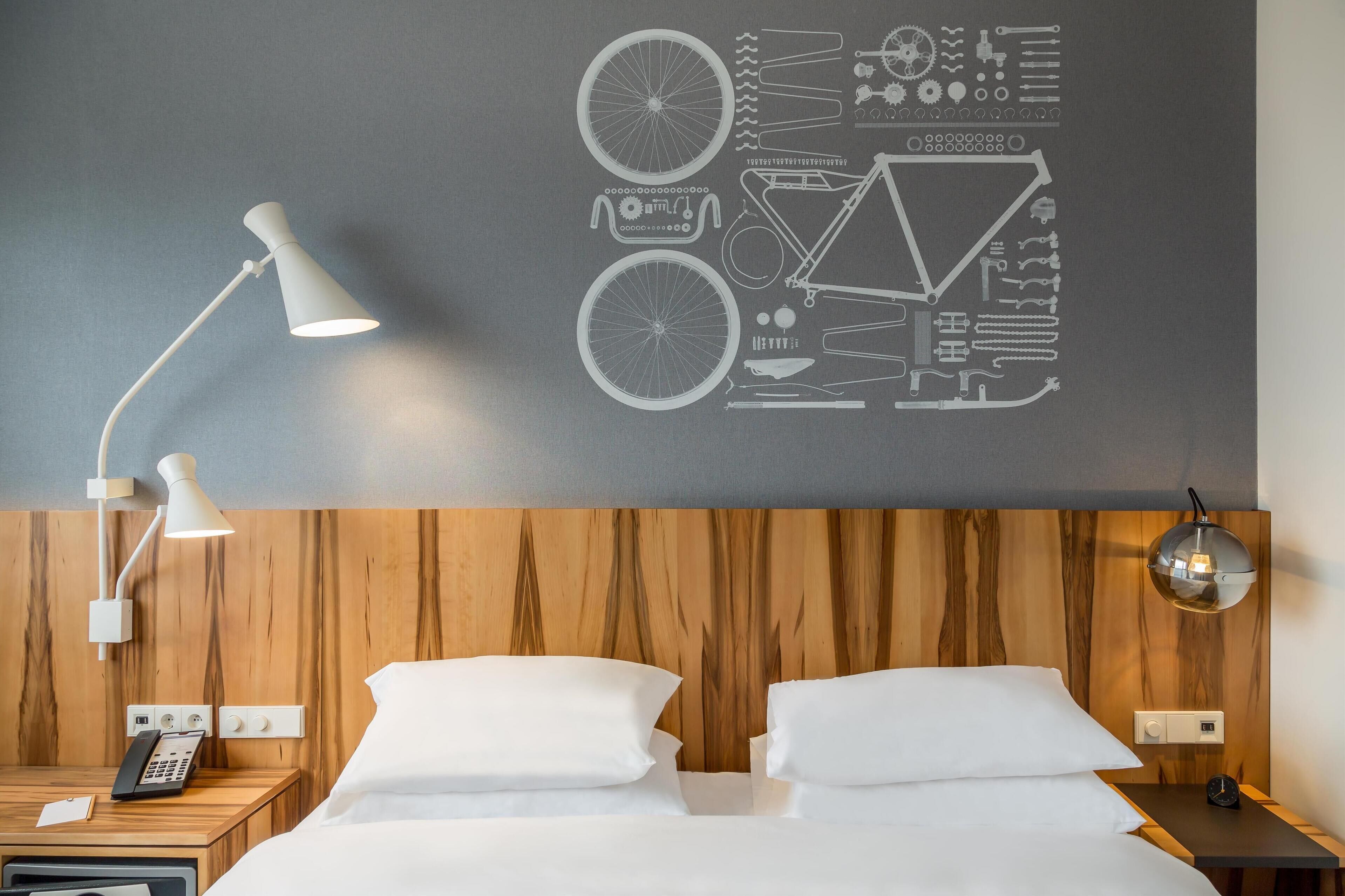 A hotel room with a wooden headboard, a bed with white linens, a bedside table with a lamp, a phone, and a wall art featuring bicycle parts.