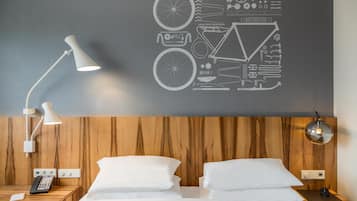 Une chambre d’hôtel avec une tête de lit en bois, un lit recouvert de draps blancs, une table de chevet avec une lampe, un téléphone et une œuvre d’art murale représentant des pièces de vélo.