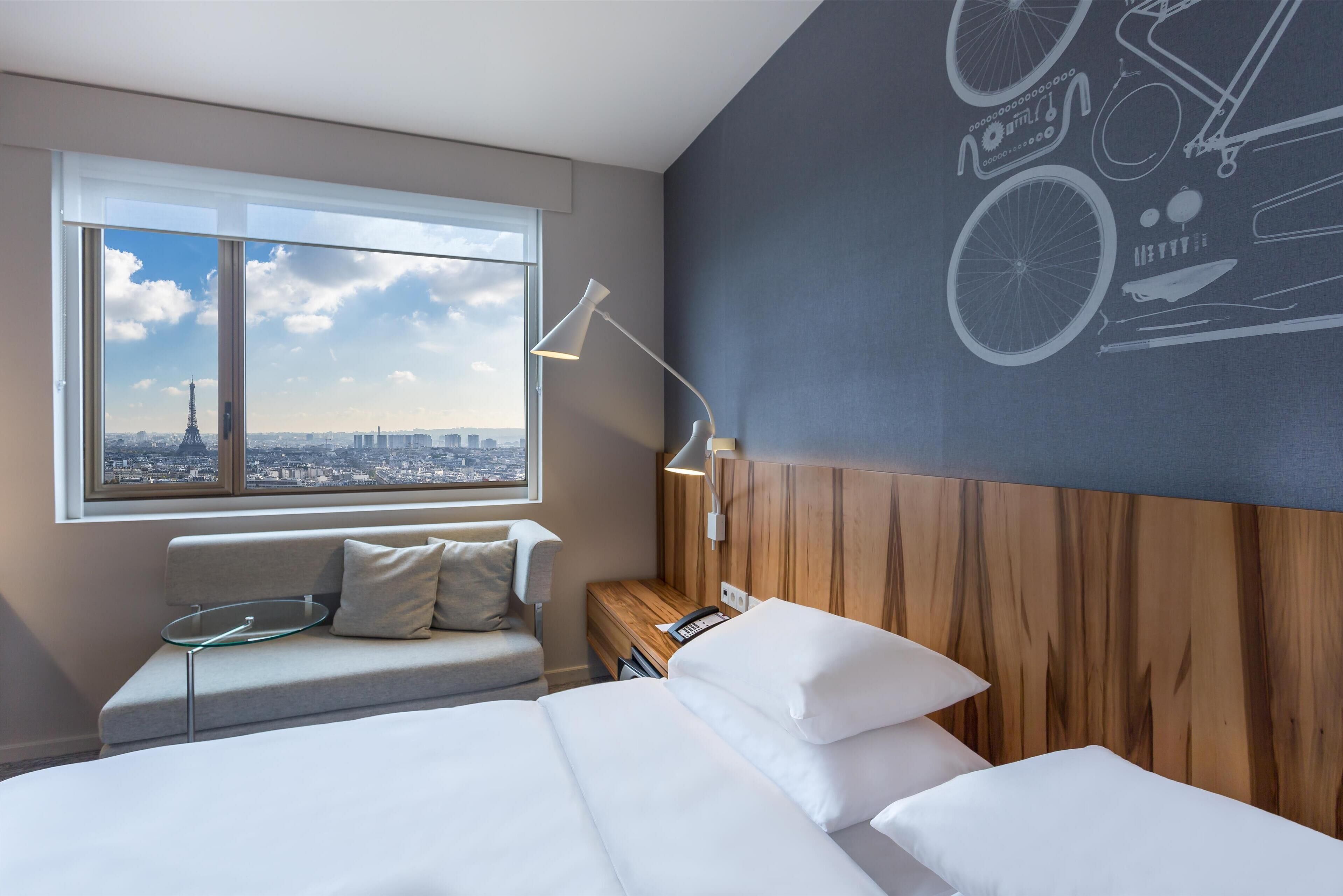 A modern hotel room with a large window offering a city view, a bed with white linens, a wooden headboard, and a bedside table with a lamp.