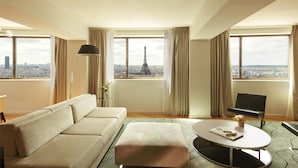 A modern hotel room with a large window offering a view of the Eiffel Tower. The room features a beige sofa, a small round table, and a black bench. - Hyatt Regency Paris Etoile (Paris)