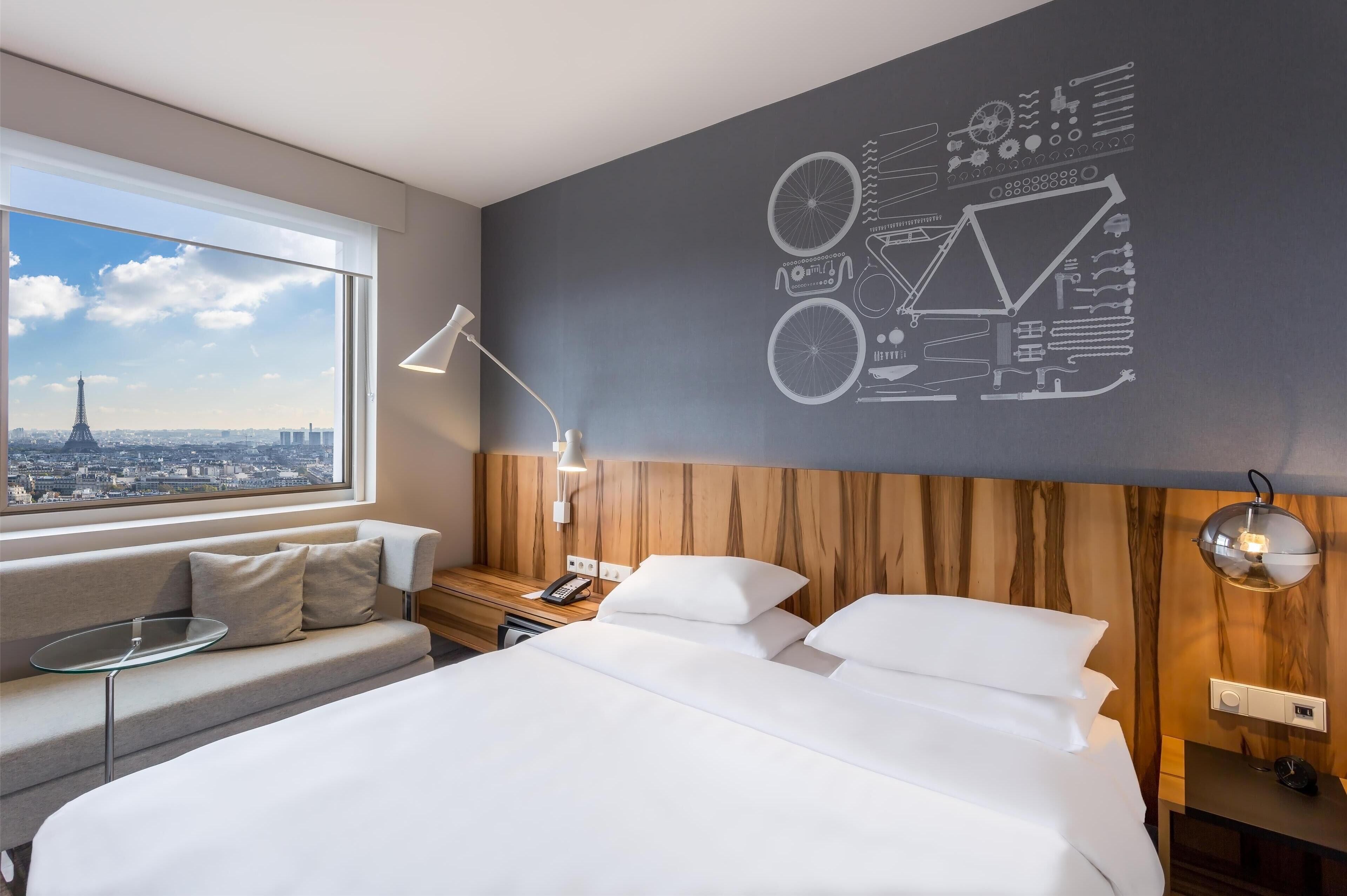 A hotel room with a bed, a sofa, a desk, a lamp, and a wall with a cityscape view.