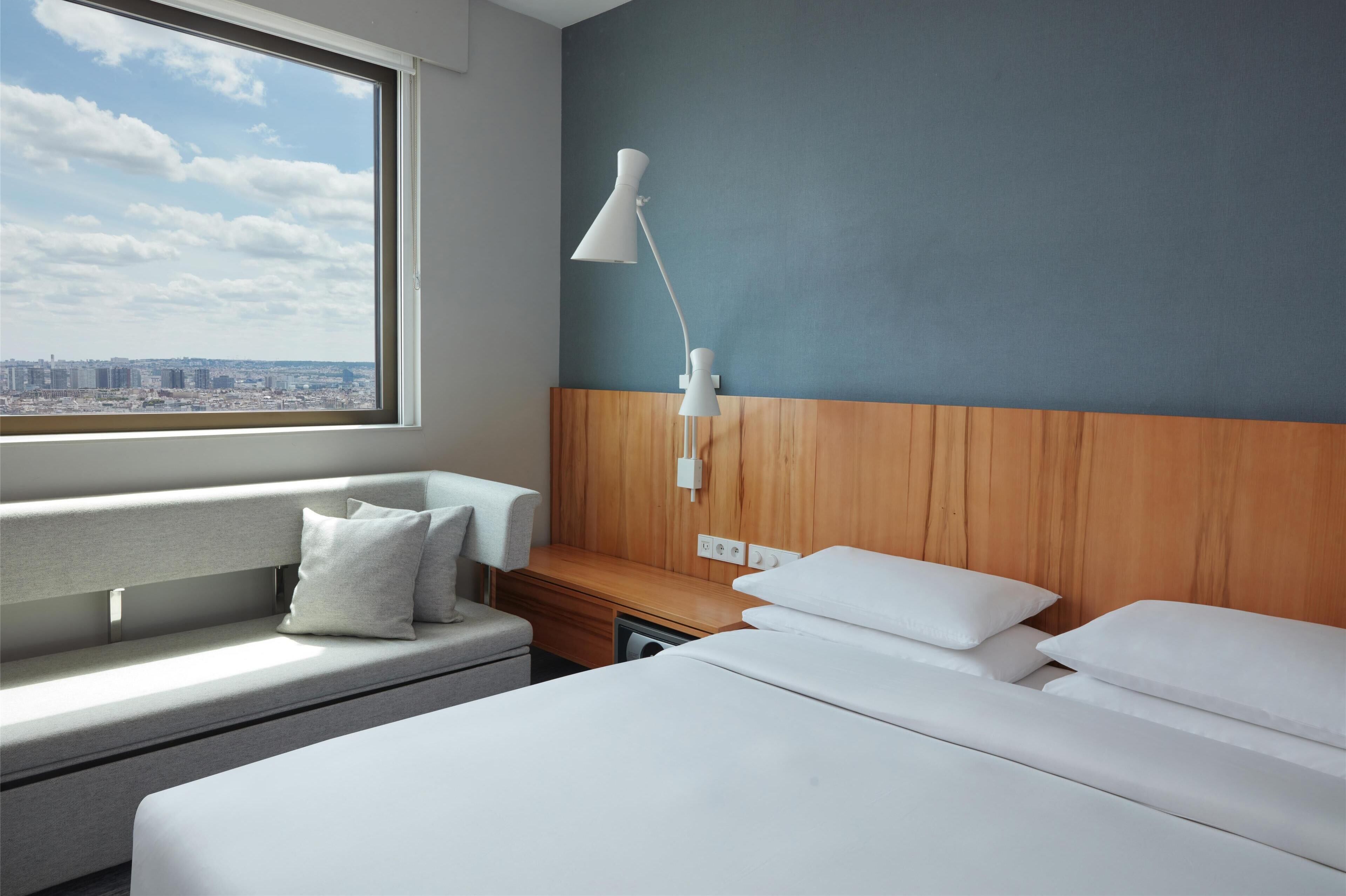 Une chambre d’hôtel avec une grande fenêtre, un lit, une table de chevet, une lampe et une vue sur la ville.