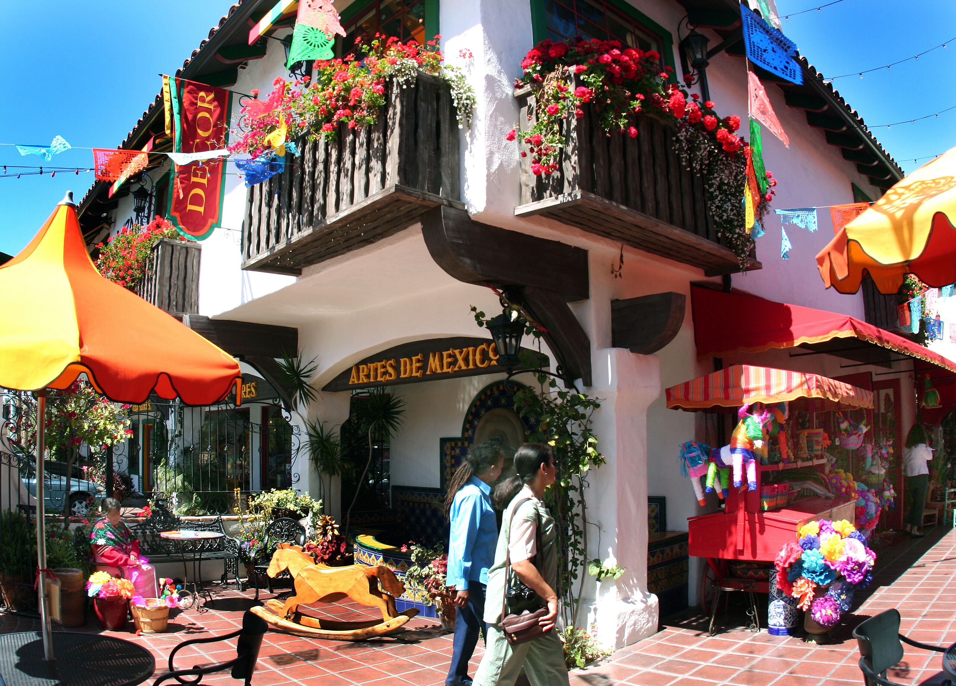 A building with a sign reading "ARTES DE MEXICO" and a balcony with flowers. There are outdoor tables and chairs, and a wooden rocking horse.