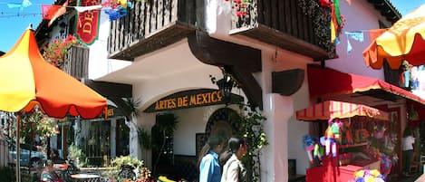 A building with a sign reading "ARTES DE MEXICO" and a balcony with flowers. There are outdoor tables and chairs, and a wooden rocking horse.
