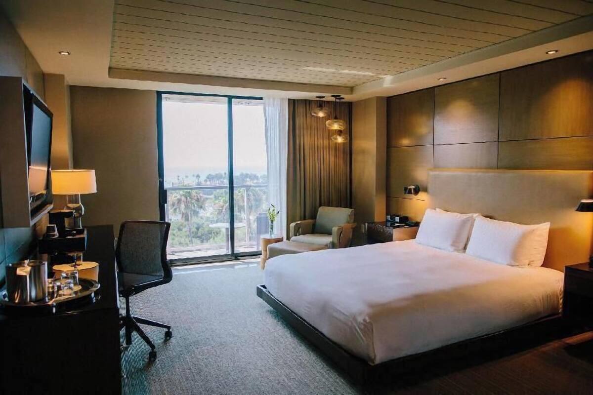 A hotel room with a large bed, a desk with a lamp, a chair, a TV, and a view of the outdoors.