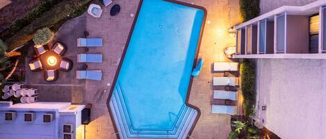 An aerial view of a swimming pool with a geometric shape, surrounded by lounge chairs and a small fire pit.