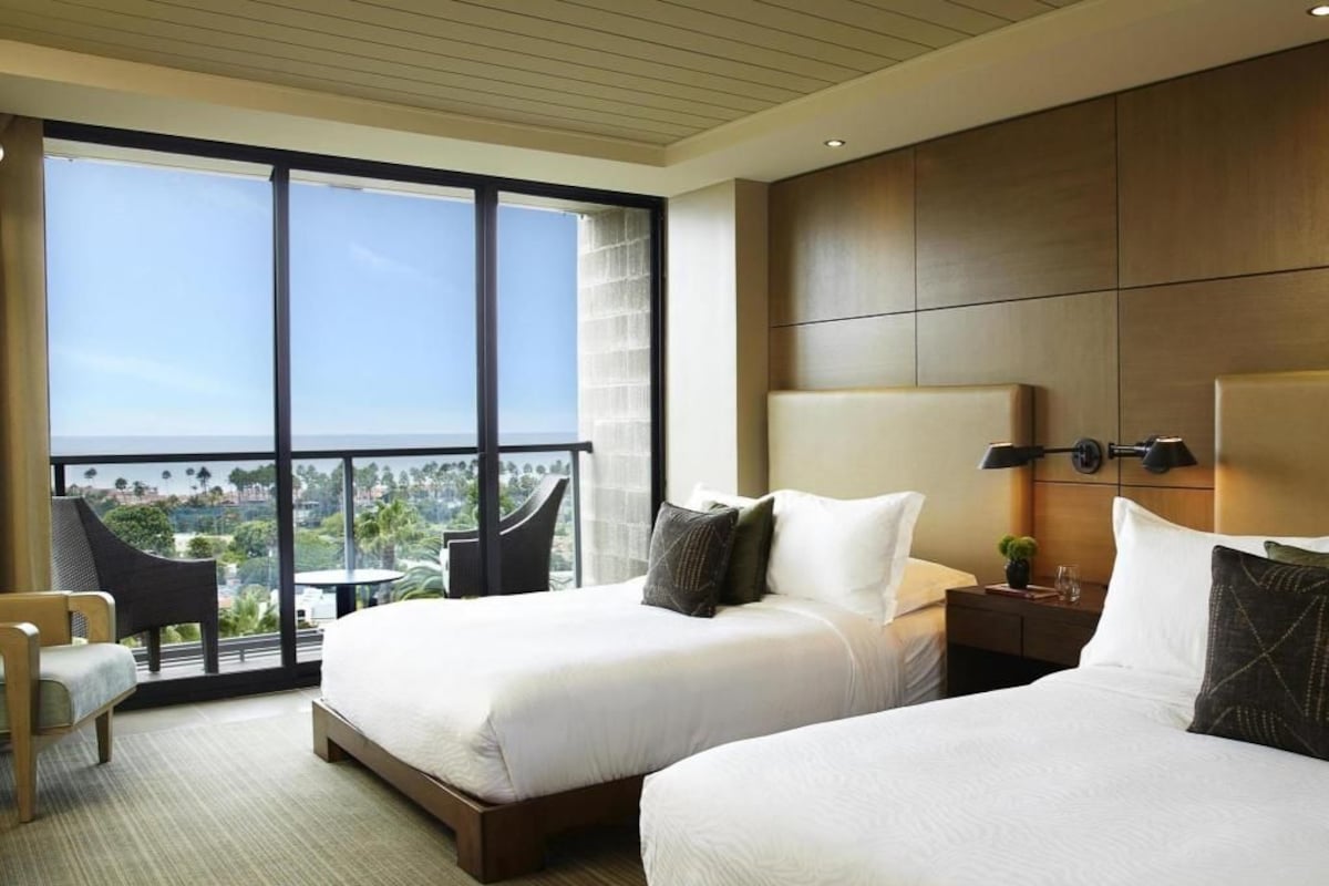 A hotel room with two beds, a view of the city, a balcony, and a chair.