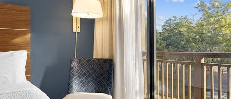 A hotel room with a bed, a nightstand, a lamp, a view of a balcony with a railing, and a window with curtains.