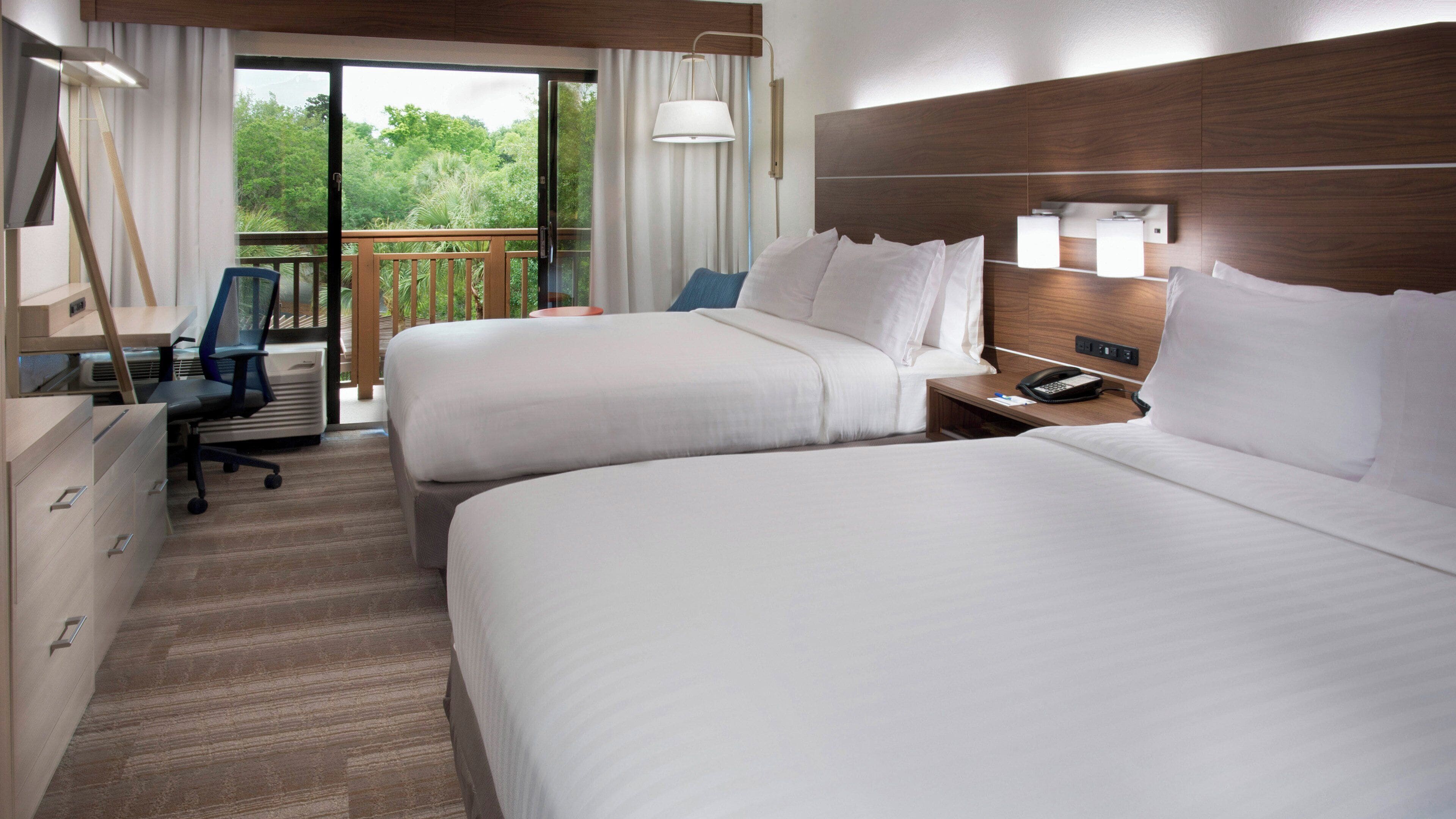 Ein Hotelzimmer mit zwei Betten, einem Schreibtisch, einem Sessel und einem Balkon mit Vorhängen.