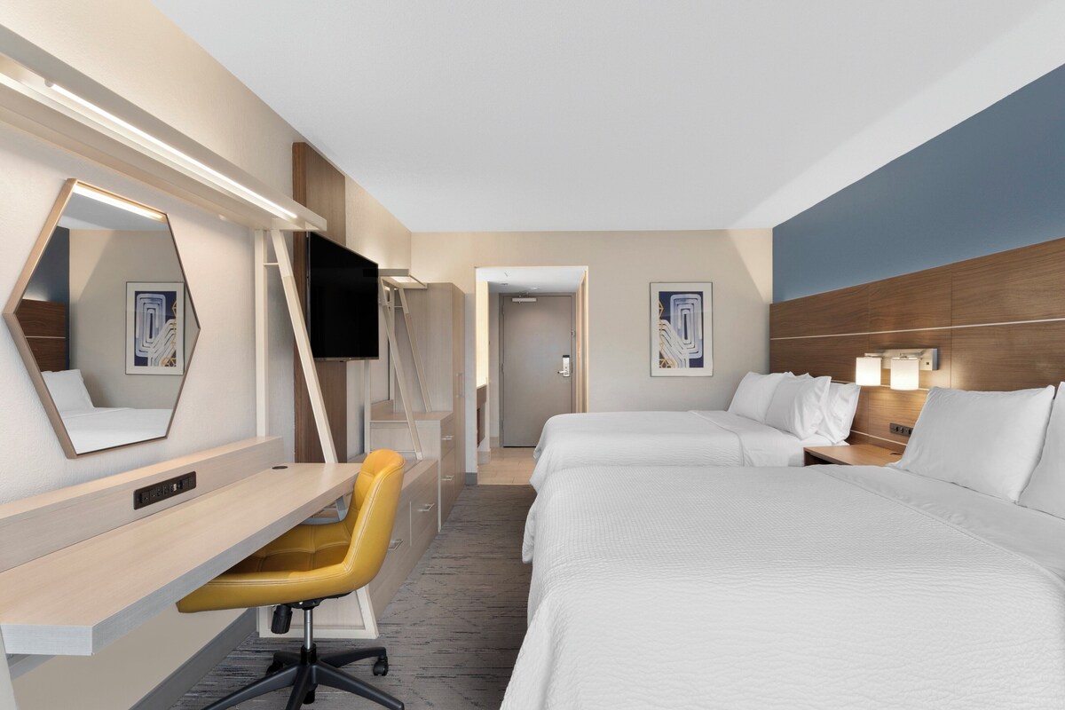 Une chambre d’hôtel équipée d’un bureau, de deux lits, d’une télévision et d’une salle de bain visible à travers une porte ouverte.