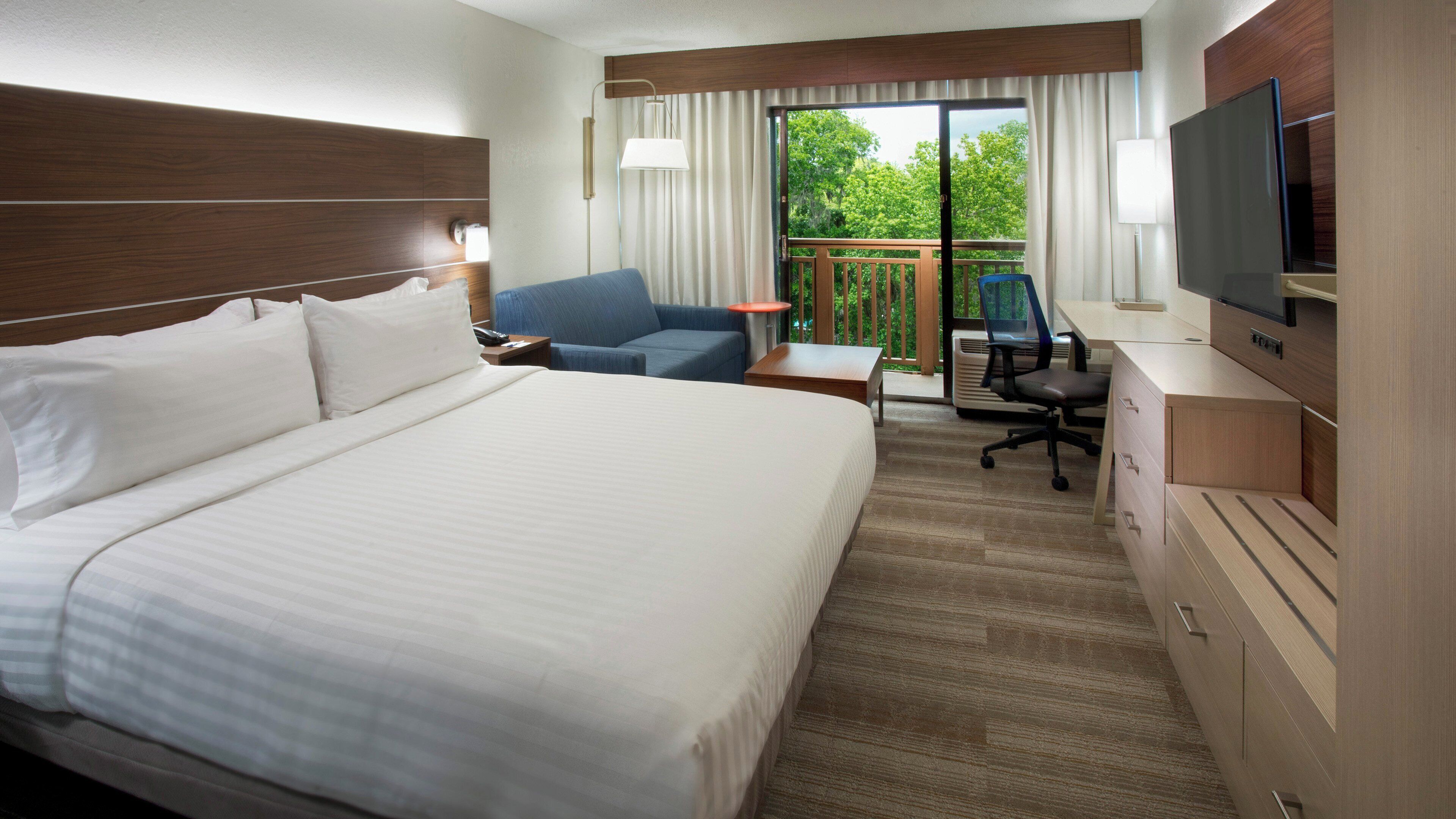 Une chambre d’hôtel avec un grand lit, un bureau avec une chaise, une télévision et un balcon donnant sur la verdure.