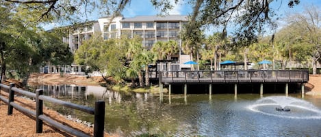 A riverside view with a wooden deck, a building with multiple floors, and a fountain in the water.