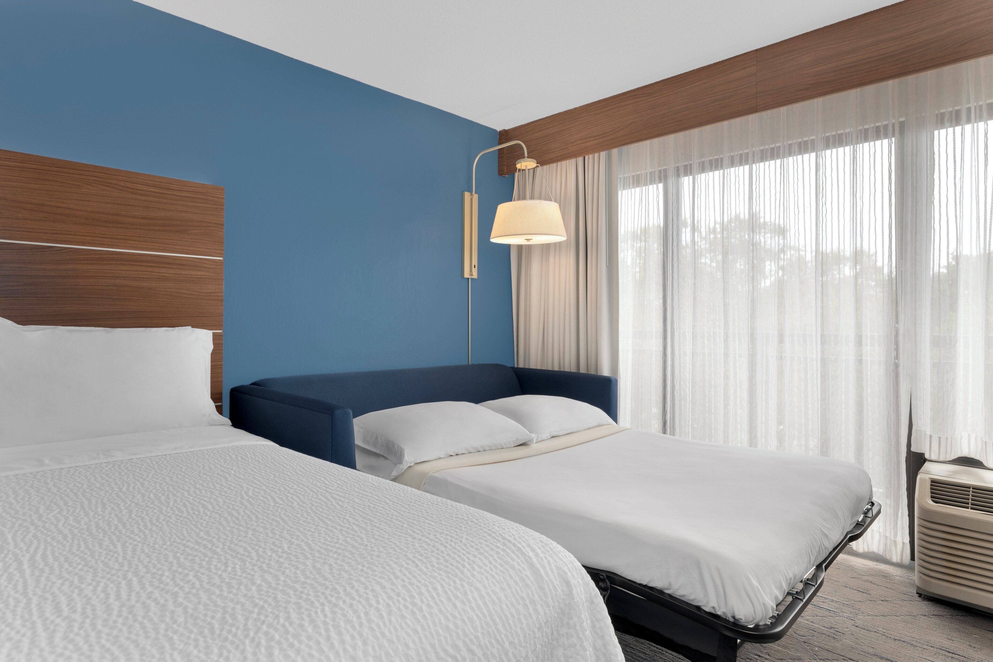Habitación de hotel con dos camas, cabecero de madera, lámpara de noche y ventana con cortinas traslúcidas.