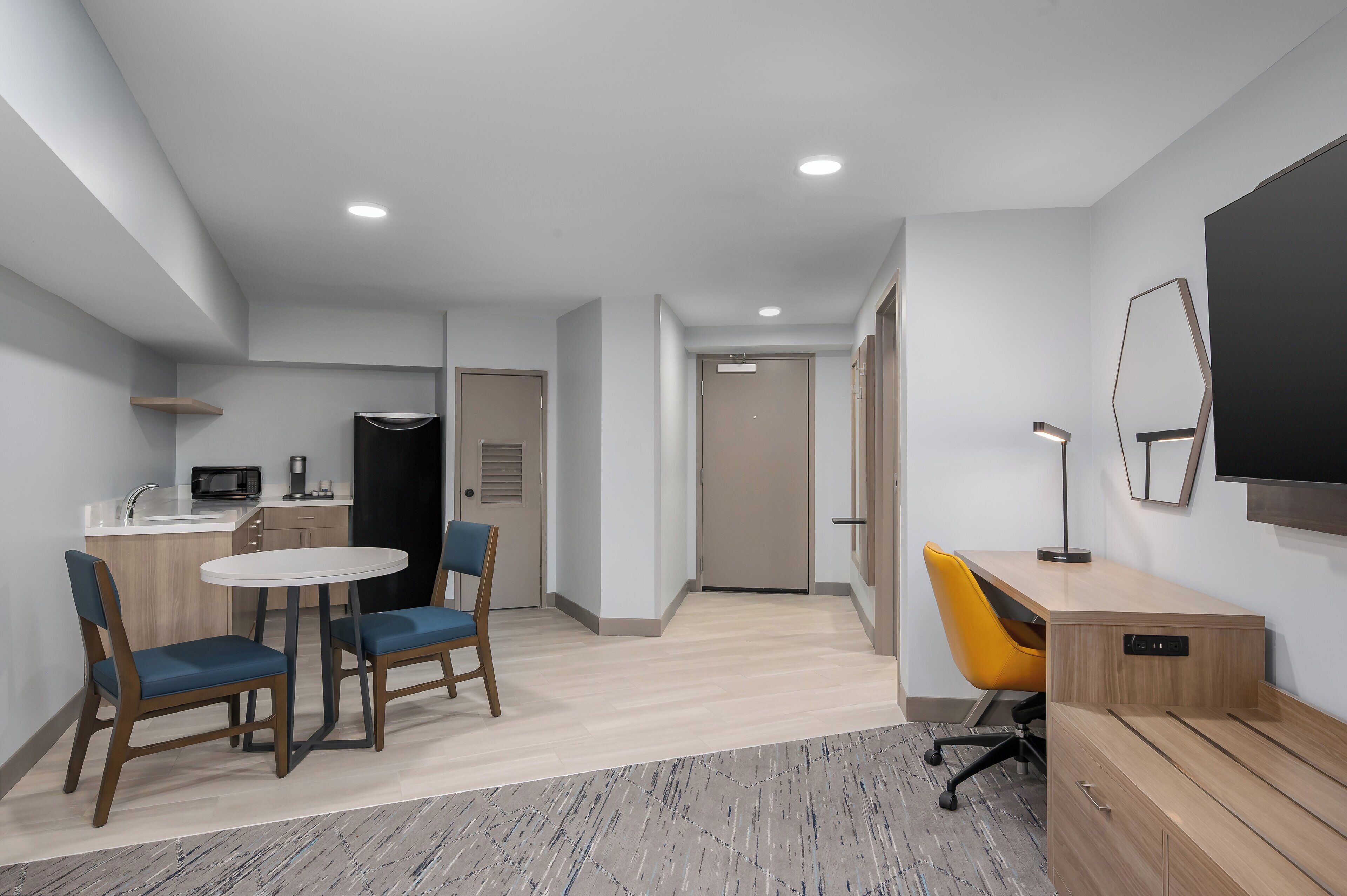 Habitación de hotel moderna con kitchenette, zona de comedor con dos sillas, escritorio y una silla, y un espejo grande.