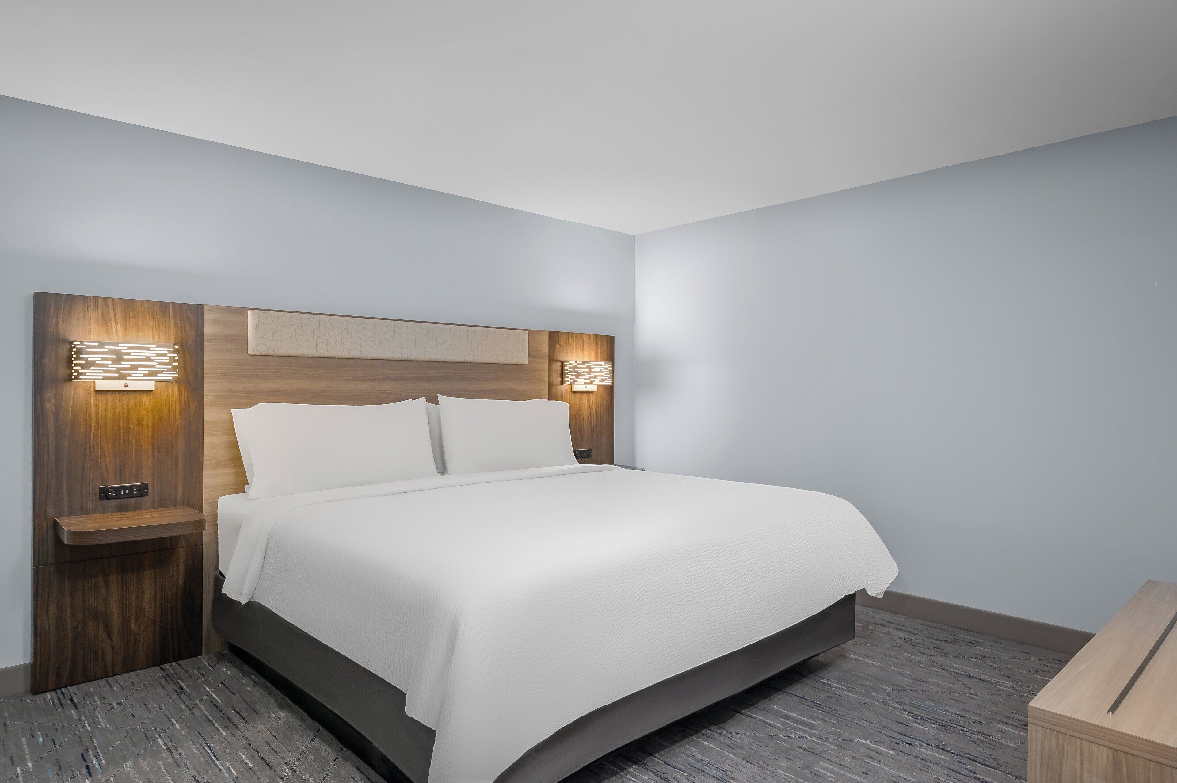 Ein modernes Hotelzimmer mit einem großen Bett, einem Holz-Kopfende und zwei Nachttischen mit integrierter Beleuchtung.