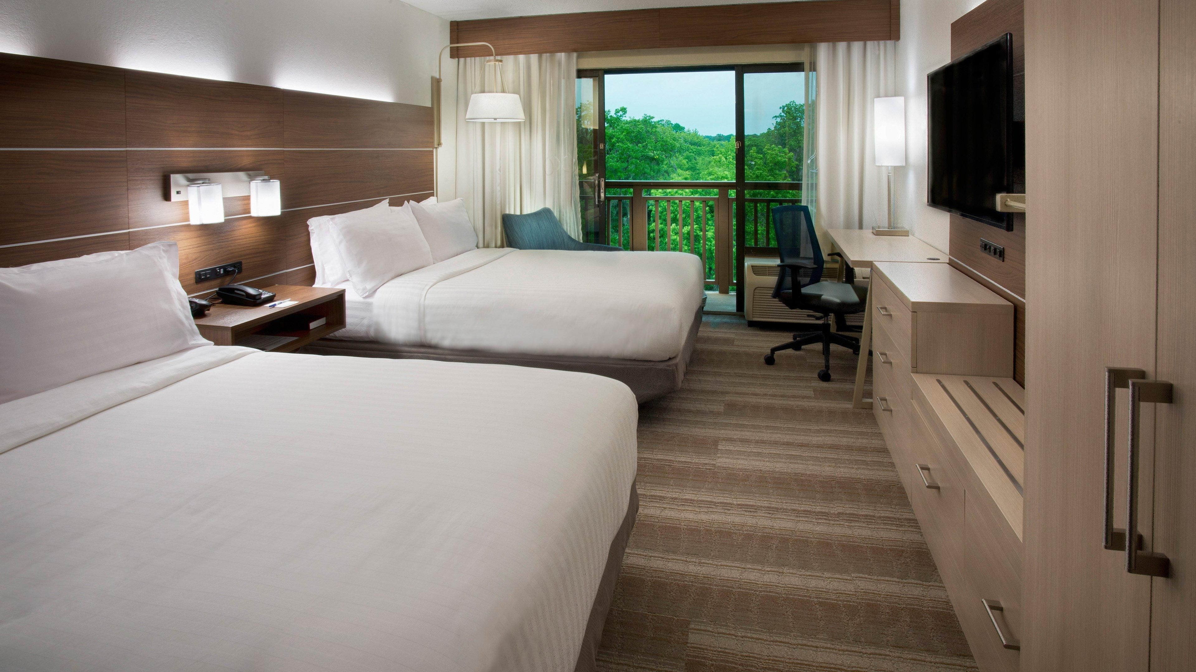 A hotel room with two beds, a desk, a chair, a TV, and a balcony with a view of greenery.