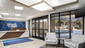 A modern reception area with a blue geometric wall, a reception desk, two grey armchairs, a small table, and a large window offering a view of the outdoors. - Holiday Inn Express Hilton Head Island by IHG (Hilton Head Island)