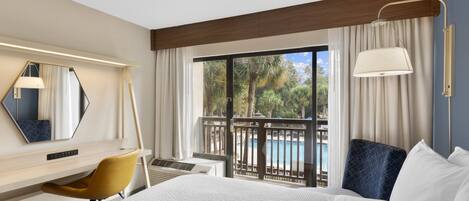 Habitación de hotel con cama, escritorio, una silla, un ventanal con vista a una piscina y palmeras, y barandilla de balcón.