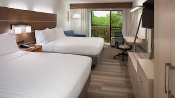 Ein Hotelzimmer mit zwei Betten, einem Schreibtisch, einem Sessel, einem Fernseher und einem Balkon mit Blick auf Grünflächen.