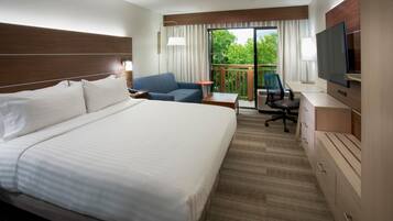 A hotel room with a large bed, a desk with a chair, a TV, and a balcony with a view of trees.