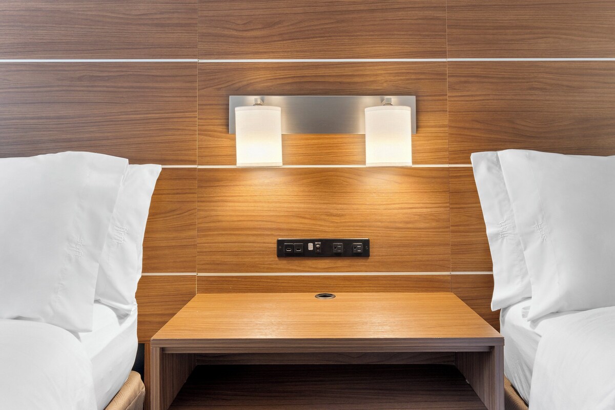 Une chambre d’hôtel avec une tête de lit en bois, deux lampes de chevet, un panneau de commande et une petite table de chevet en bois.
