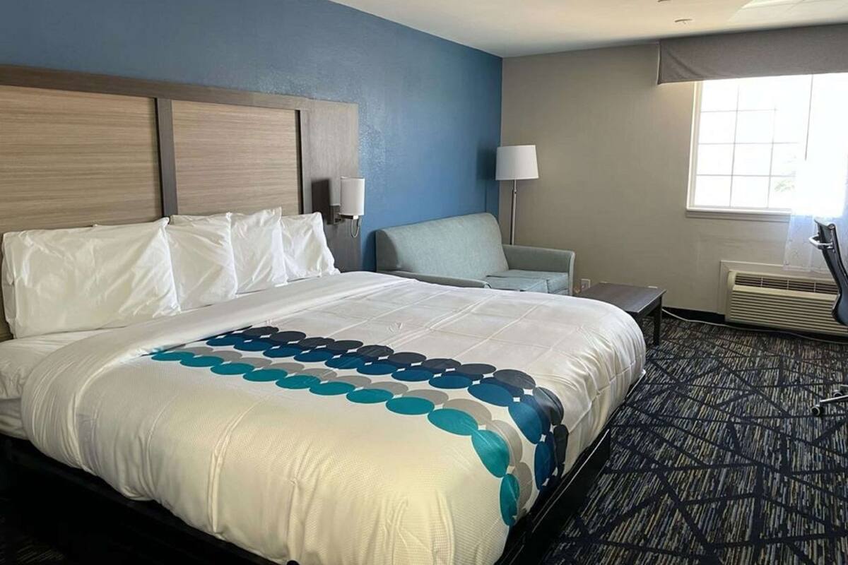A hotel room with a large bed, a chair, a desk, a lamp, and an air conditioning unit.
