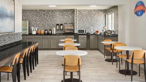 A modern kitchen with a black and white patterned backsplash, a microwave, and a coffee machine. There are multiple round tables and chairs arranged for dining.
