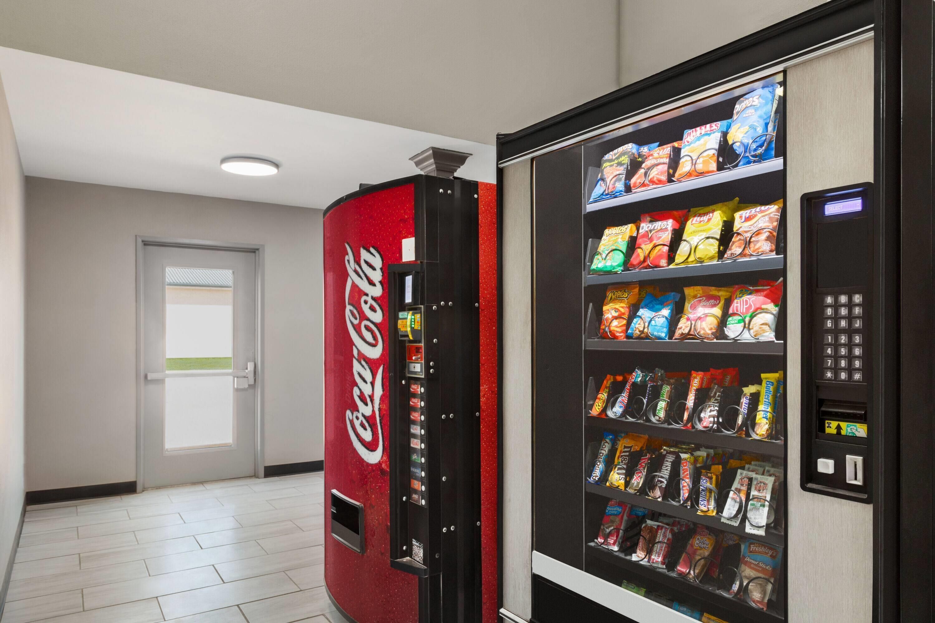 En automattillbehörsmaskin med snacks och en läskautomat med Coca-Cola-produkter, placerade i en korridor.