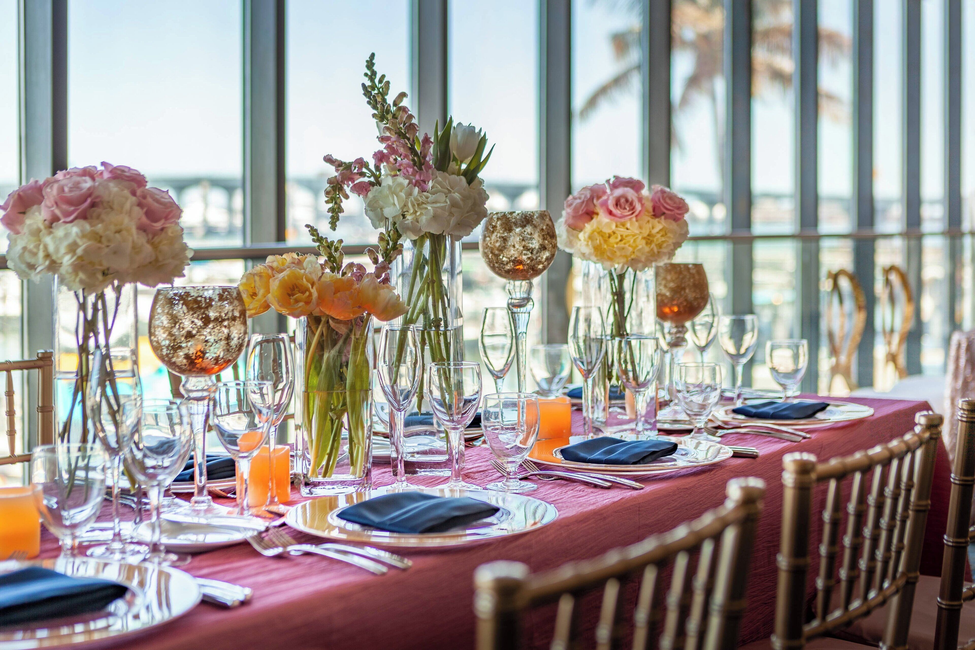 Una mesa dispuesta para una comida, con centros de mesa florales, cristalería transparente y un mantel rojo.