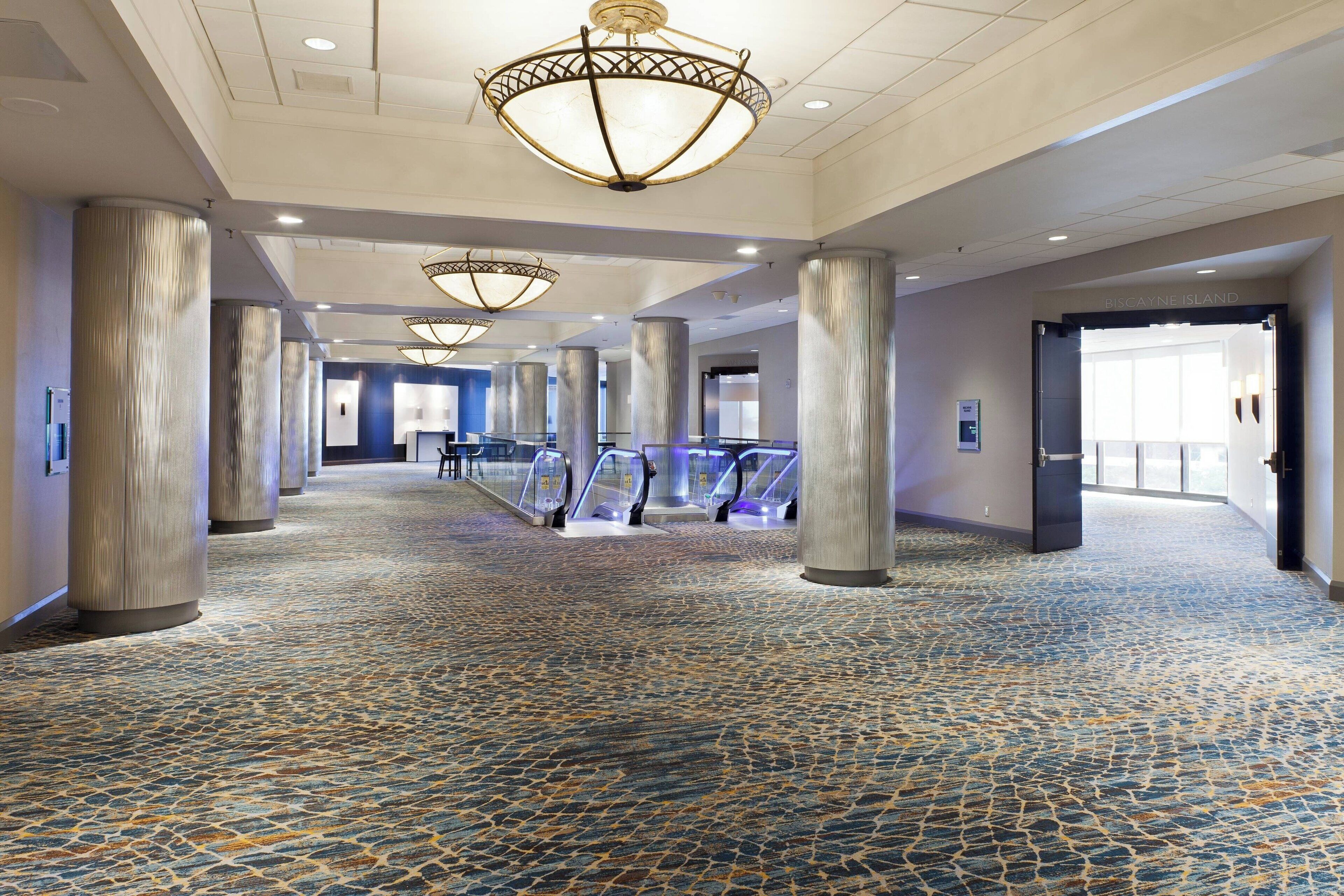 A spacious hallway with a patterned carpet, large columns, and modern lighting fixtures.