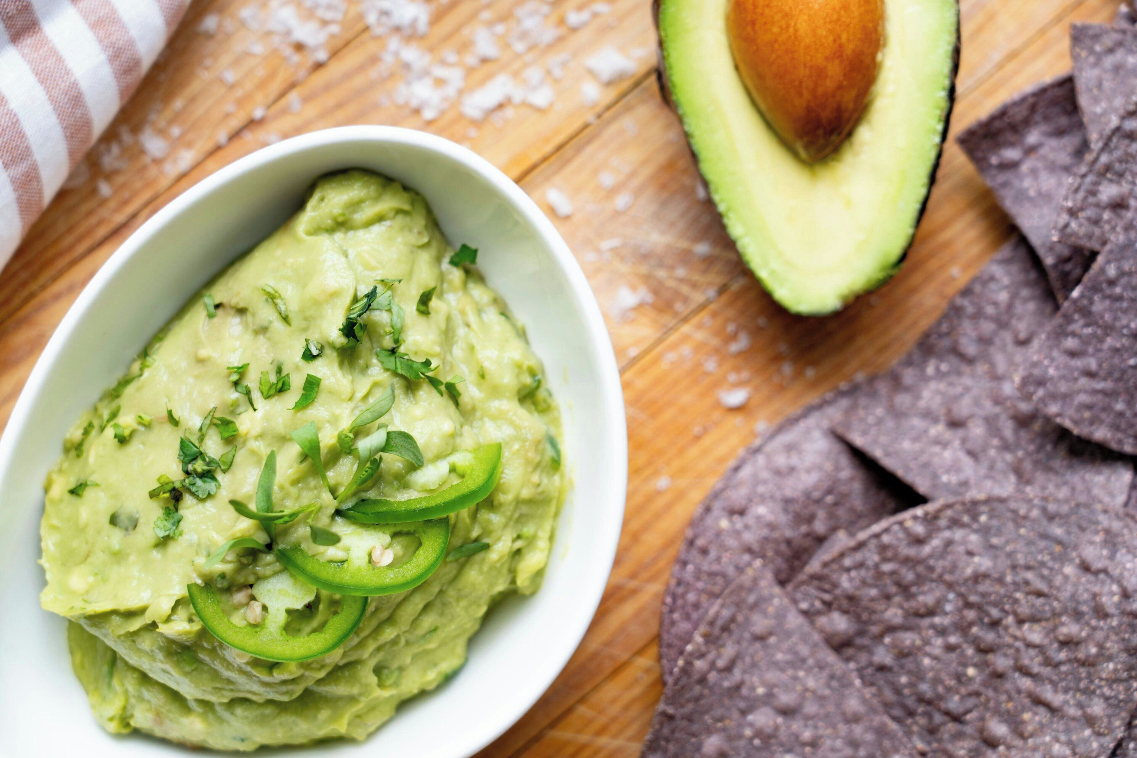 Eine Schüssel Guacamole mit geschnittenen Jalapeños und gehackten Kräutern, umgeben von Tortilla-Chips und einer halbierten Avocado auf einer Holztischplatte.