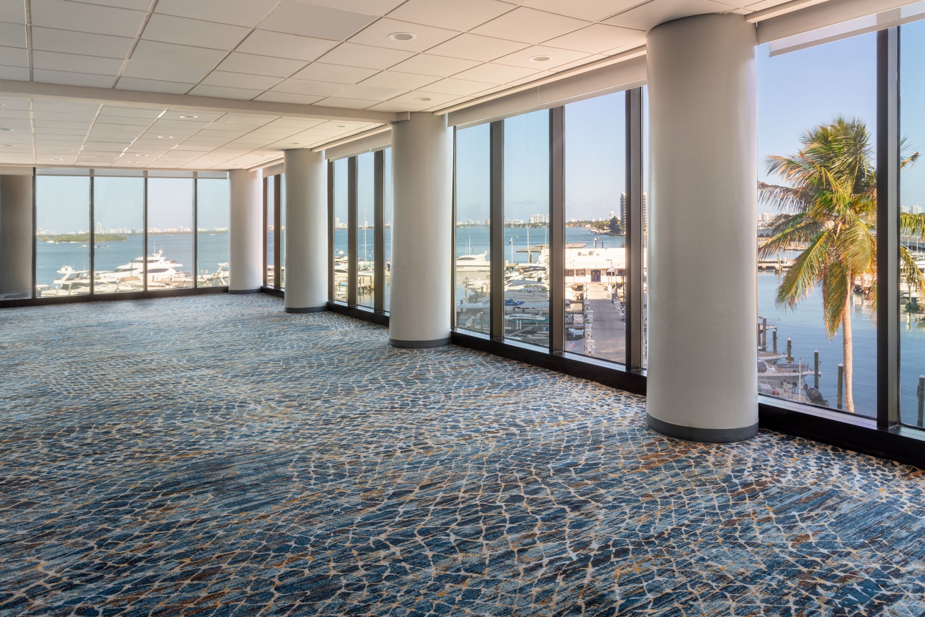 A spacious indoor area with large windows offering views of a marina and palm trees.